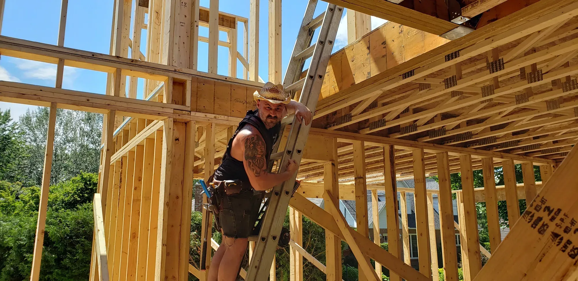 Maxime Roy Bouliane, chapeau de paille sur la tête, appuyé à une échelle au milieu de l'ossature de bois d'une maison neuve