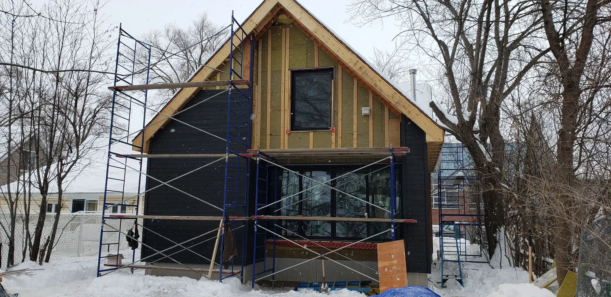 Pignon d'une maison en cours de revêtement l'hiver : moitié en planches de bois noir posées, moitié encore en isolant nu, échafaudage monté